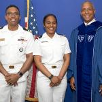 President Riley in Commencement robe standing with Sierrah Grigsby in dress whites along with another naval officer in dress whites.